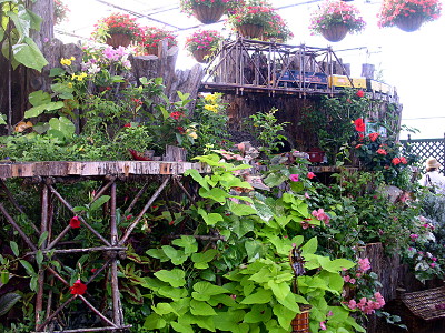 [More than a half-dozen potted plants with red blooms hang from the ceiling. There are a multitude of different colored blooming plants as well as a plant in the foreground which only has bright large light-green leaves. A worn rusty-looking structure supports different levels of train tracks as the train weaves around the plants.]